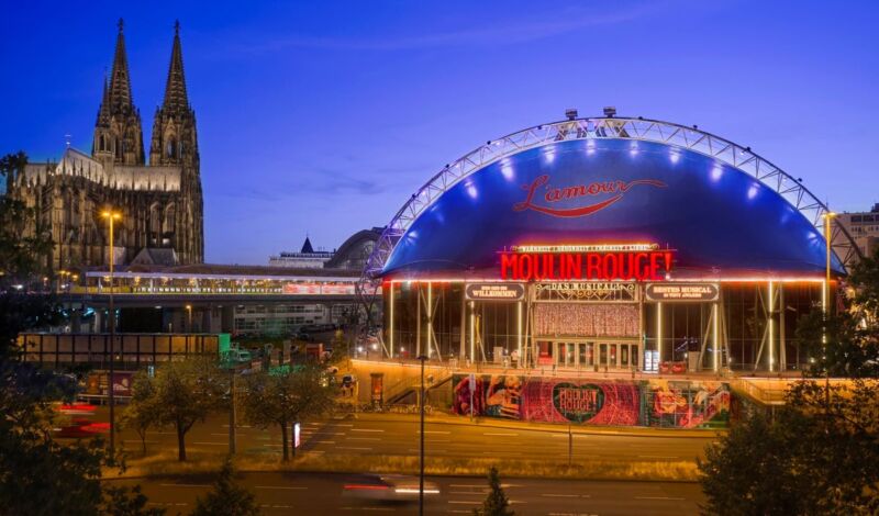 Die Außenansicht des Musical Domes in Köln mit dem Schriftzug des Musicals "Moulin Rouge". Im Hintergrund ist der Kölner Dom zu sehen,