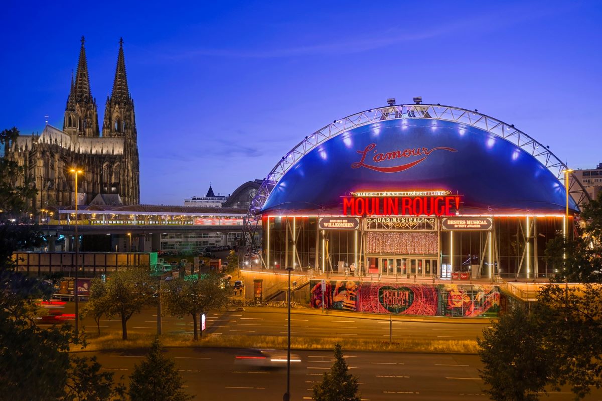 Die Außenansicht des Musical Domes in Köln mit dem Schriftzug des Musicals "Moulin Rouge". Im Hintergrund ist der Kölner Dom zu sehen,