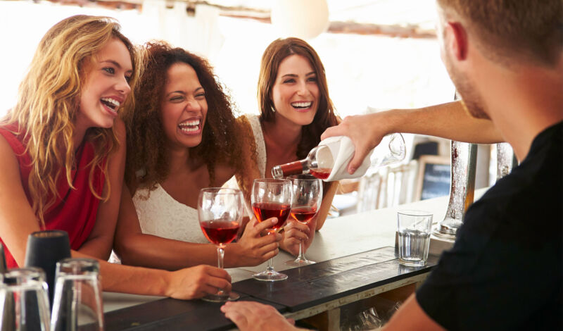 Drei junge Frauen lassen sich an der Theke Wein einschenken.