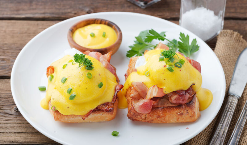 Eggs Benedict auf einem Teller, dazu Sauce Hollandaise und Besteck