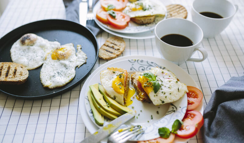 Ein gedekcter Frühstückstisch unter anderem mit Spiegelei, Avocado und schwarzem Kaffee.