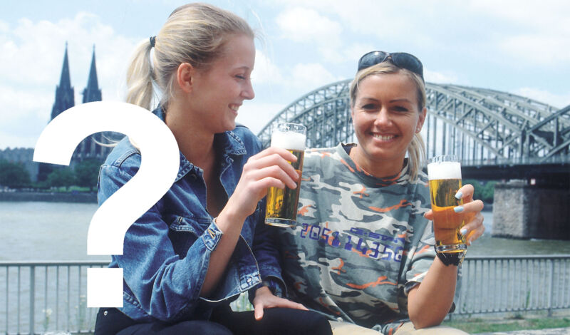 Zwei Frauen stoßen mit Kölsch an, im Hintergrund die Hohenzollenrbrücke und der Kölner Dom, über dem Foto liegt ein Fragezeichen