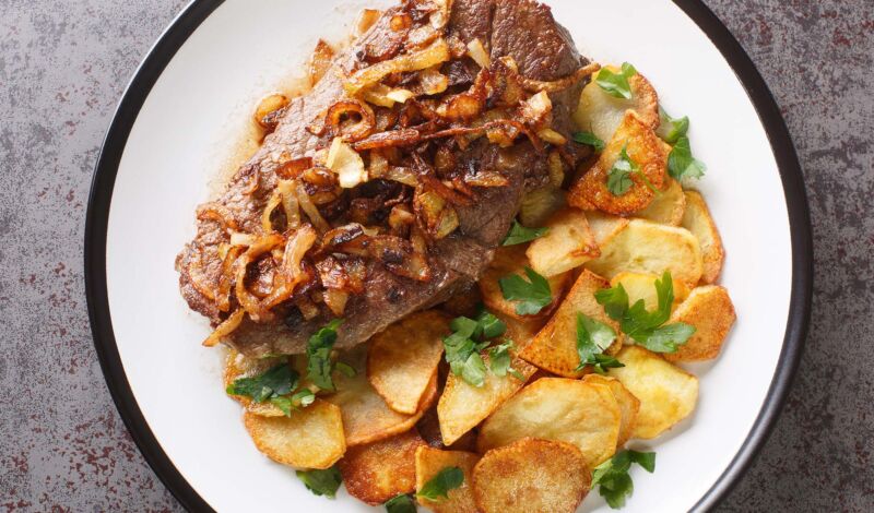 Ein Steak mit Zwiebeln, angerichtet auf einem weißen teller mit Bratkartoffeln.