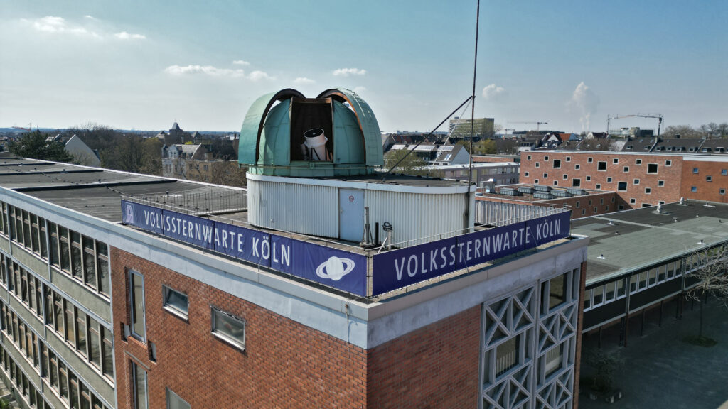 Ein Blick von Außen auf das Dach der Volkssternwarte Köln. Auf dem Dach ist eine Kuppel geöffnet, aus der ein Teleskop Lugt.