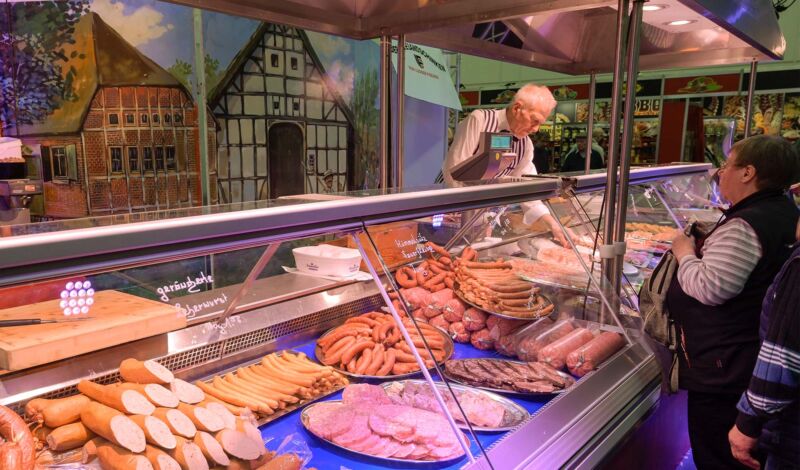 Kunden stehen an an einer Theke in einer Metzgerei, die mit Fleisch- und Wurstwaren gefüllt ist.