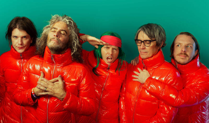 Die Mitglieder der Rockband The Flaming Lips gekleidet in rote Steppjacken vor einem grünen hintergrund