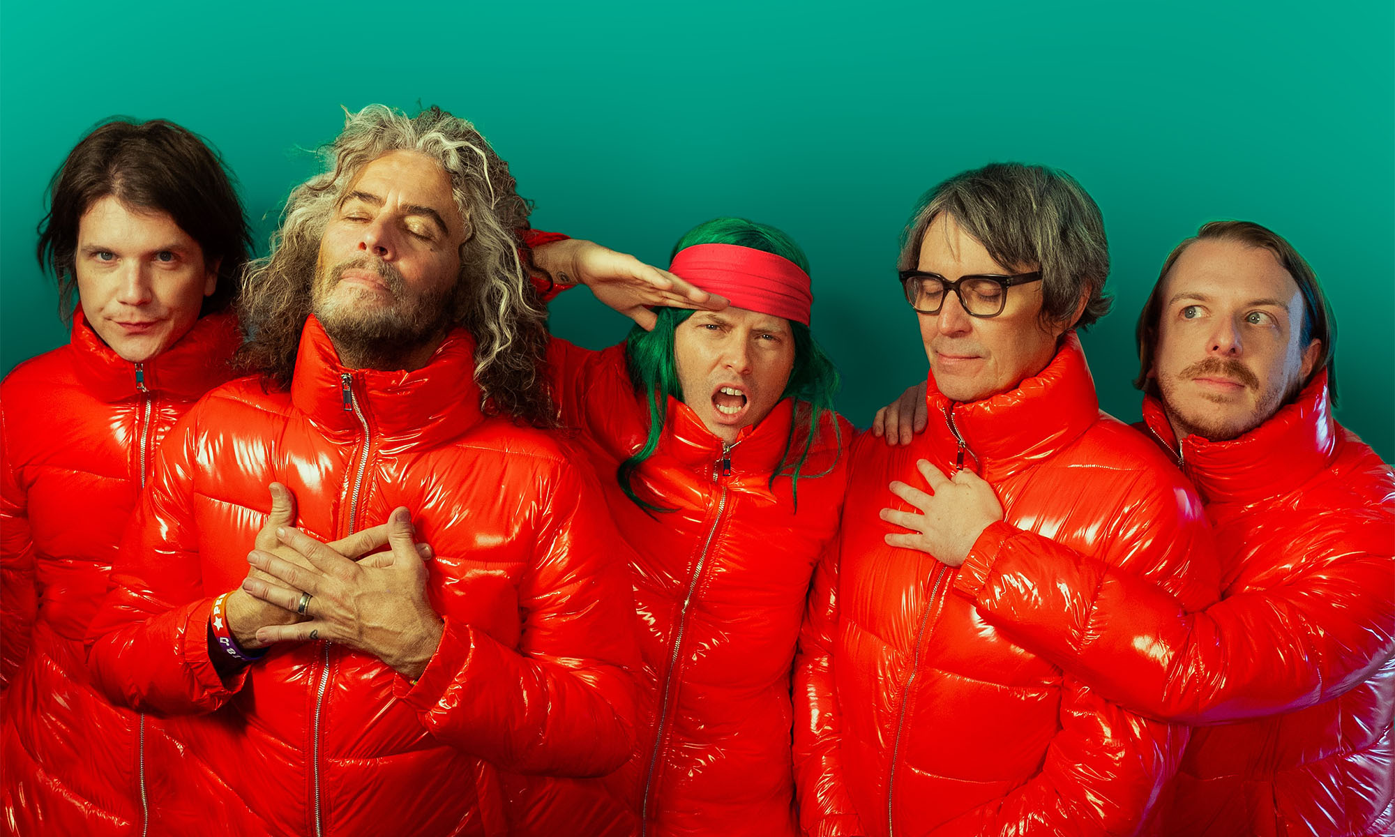 Die Mitglieder der Rockband The Flaming Lips gekleidet in rote Steppjacken vor einem grünen hintergrund