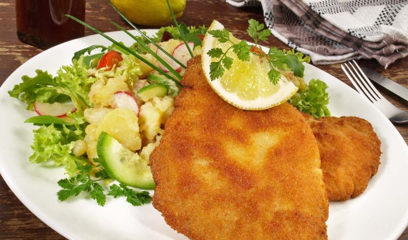 Wiener Schnitzel mit Kartoffesalat und Gurkensalat angerichtet auf einem weißen Teller.