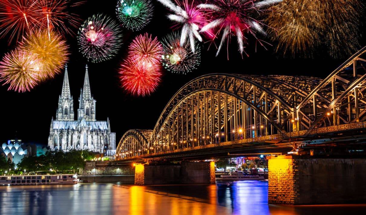 Das Foto zeigt einen Blick auf den Kölner Dom und die Hohenzollernbrücke in der Silvesternacht mit explodierendem Feuerwerk am Nachthimmel.